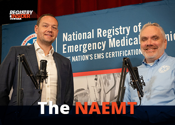 Two men stand in front of a backdrop featuring the National Registry of Emergency Medical Technicians, with microphones set up for a discussion.